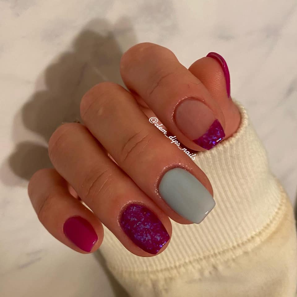 Hand with nails painted in matte dusty blue, glossy magenta, and glittery purple accents on a marble background.