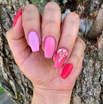 Hand with pink and coral matte nails, one accent nail decorated with gold flakes on ring finger.