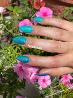 Teal blue glittery dip powder nails with multicolor chameleon flakes on hand over pink flowers outdoors.