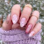 Fingernails with pink and red glittery chrome dip powder polish in almond shape on hand with gold rings and purple knit.