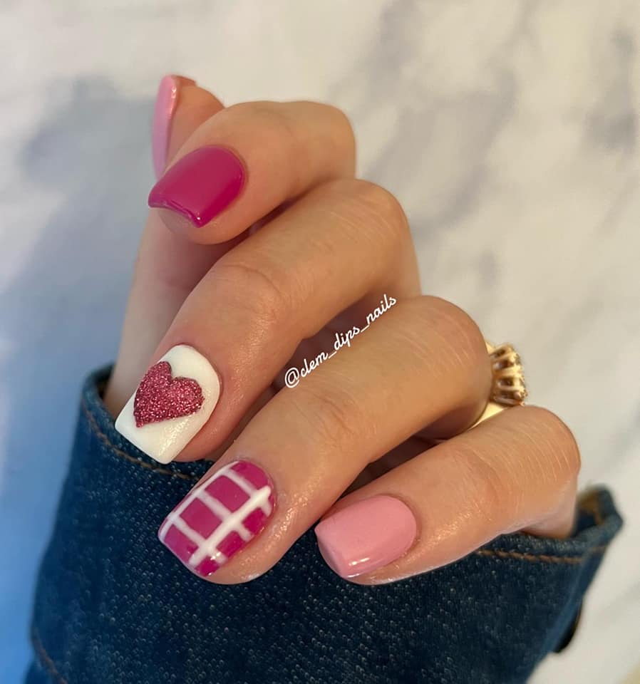 Manicured hand with hot pink dip powder nails, featuring a white accent nail with a pink glitter heart and a pink and white.