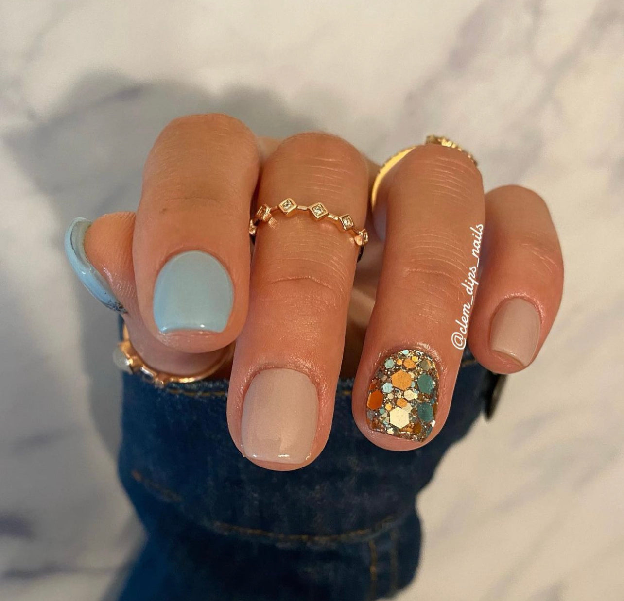 Hand with short nails showing beige, nude, light blue, and glittery turquoise nail polish with gold accents.
