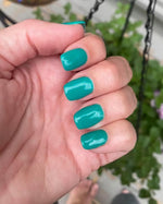 Teal green glossy dip powder polish on short square nails against blurred plants background.
