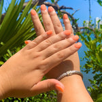 Hands with pastel coral orange dip powder nails against a sunny blue sky and green palm leaves background.