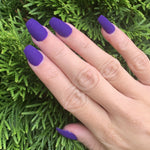 Matte purple coffin-shaped nails on hand against green foliage background.
