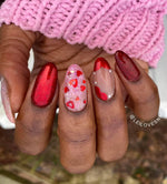 Hand with acrylic nails in red chrome, red glitter, glossy red tips, and nude with red hearts design.