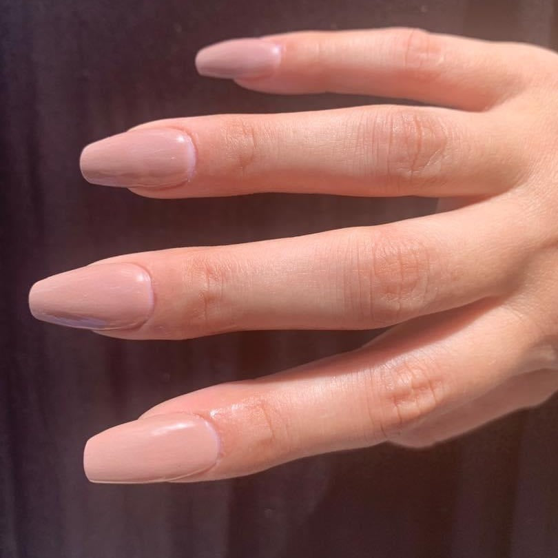 Long almond-shaped nails with glossy nude tan dip powder polish on a hand against a dark background.