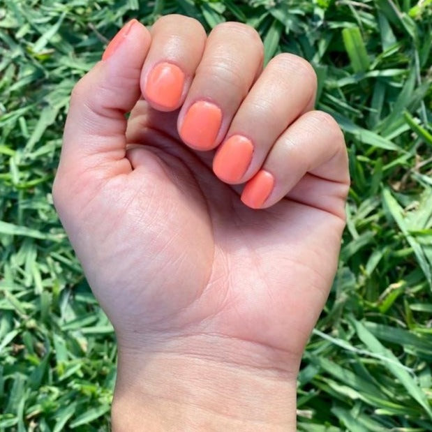 Hand with short nails painted in solid peach-orange dip powder polish against green grass background.