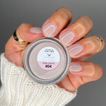 Hand holding small jar of beige dip powder nail polish with matching nude polished nails and gold rings.
