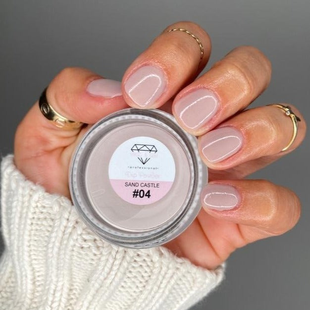 Hand holding small jar of beige dip powder nail polish with matching nude polished nails and gold rings.